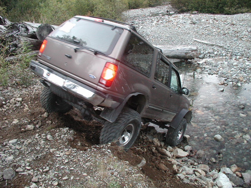 Brown SUV driving through a rocky stream and up a dirt embankment.