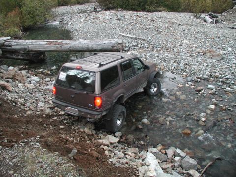 1985 Ford Bronco - A brown SUV is navigating a rocky stream bed, with a large fallen log spanning the water in the background. The vehicle appears to be modif…