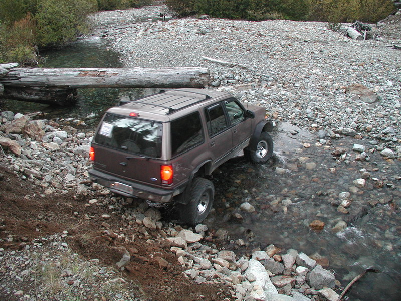 Brown SUV driving through a rocky stream with a large log bridge in the background.