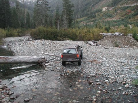 1985 Ford Bronco - A brown SUV is parked on a rocky riverbed, with a large log bridging the water in the foreground. The vehicle is facing away from the camer…