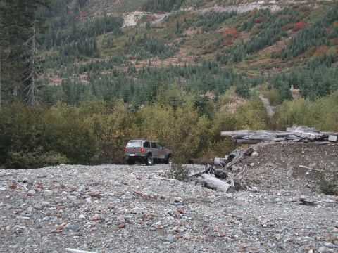 1985 Ford Bronco - A brown SUV is parked on a rocky, gravelly terrain with a dense forest and a hillside in the background. The vehicle is viewed from the rea…