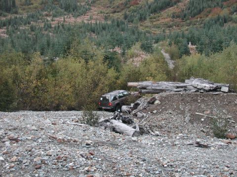 1985 Ford Bronco - A dark colored SUV is parked on a rocky, debris-strewn slope next to a pile of logs and branches. The vehicle is surrounded by trees and de…