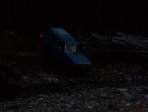 1985 Ford Bronco - A blue pickup truck with a camper shell is parked at a steep angle on a rocky, wooded embankment next to a stream.