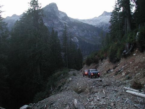 1985 Ford Bronco - Two blue pickup trucks drive down a rough, rocky mountain road surrounded by dense evergreen trees. The lead truck is a dark blue pickup wi…