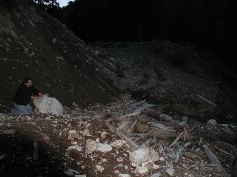 1985 Ford Bronco - A man kneels on the ground next to a large rock on a dark, debris-strewn hillside. The scene appears to be a landslide area with trees visi…