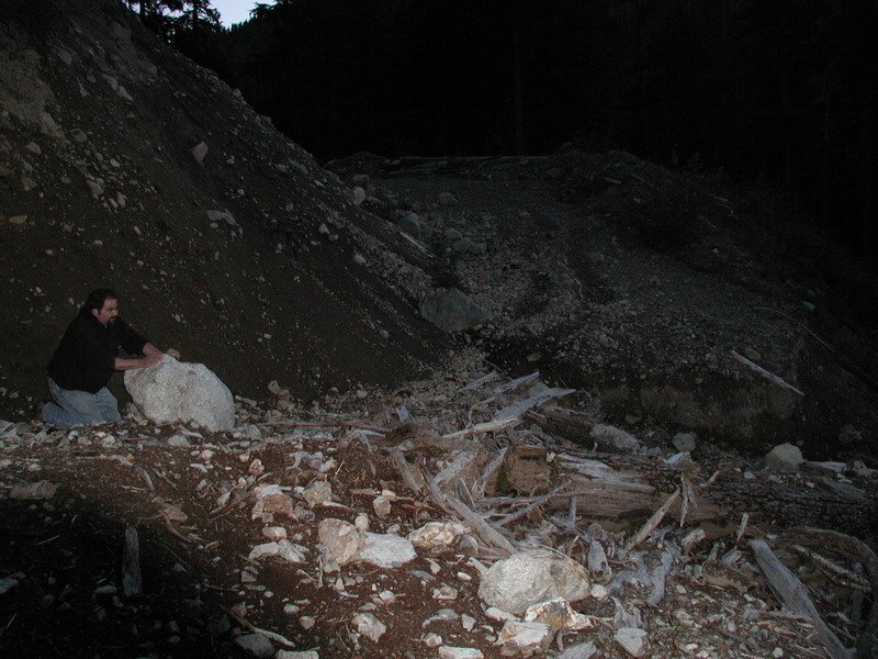 Man kneeling by a large rock on a dark, debris-strewn hillside.