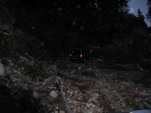 1985 Ford Bronco - A dark-colored SUV is partially visible on a steep, rocky, and wooded incline at night. The vehicle's taillights are illuminated, suggestin…