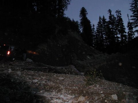 1985 Ford Bronco - A dark scene shows a dark-colored SUV parked on a rocky, wooded trail at dusk. The vehicle's taillights are illuminated, and a person stand…