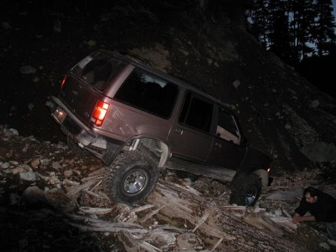 1985 Ford Bronco - A dark-colored SUV is angled precariously on a steep, rocky, and debris-strewn incline at night. A person is visible in the lower right cor…