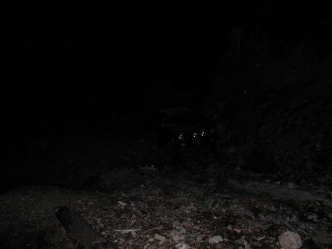 1985 Ford Bronco - A dark colored Ford Bronco is parked on a rocky, uneven terrain at night. The headlights of the vehicle are illuminated.