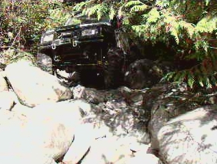 1985 Ford Bronco - A black 1985 Ford Bronco is navigating a rocky, wooded trail, with its front tire visible on the right side of the frame.