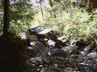 1985 Ford Bronco - A white 1985 Ford Bronco is navigating a rocky, wooded stream bed. The vehicle is angled towards the viewer's left, with its front end slig…
