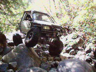 1985 Ford Bronco - A modified white off-road vehicle navigates a rocky, wooded trail, with its front wheels lifted high off the ground.