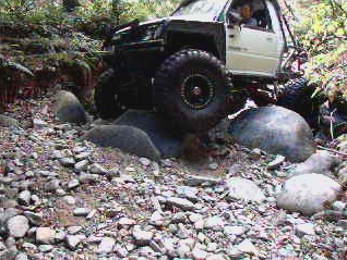 1985 Ford Bronco - A white off-road vehicle navigates a rocky, wooded trail, with its large tires and suspension clearly visible. The vehicle is angled toward…