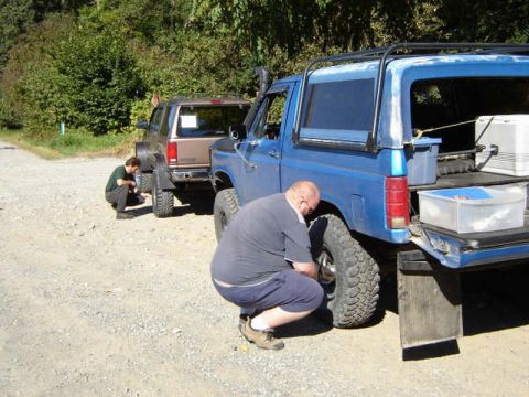 1985 Ford Bronco - Two men are working on the tires of two off-road vehicles parked on a gravel road. One man is squatting by the rear tire of a blue Ford Bro…