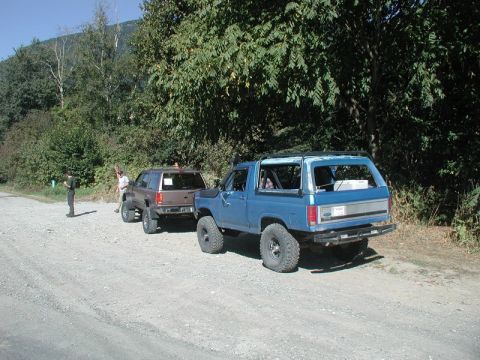 1985 Ford Bronco - A blue 1985 Ford Bronco with its top removed is parked on a gravel road next to a brown SUV. The Bronco has large off-road tires and a cust…