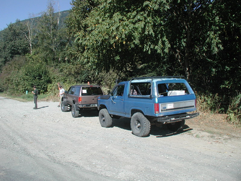 Blue 1985 Ford Bronco with top removed, parked on gravel road next to brown SUV.