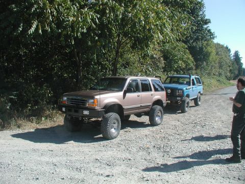 1985 Ford Bronco - Two lifted off-road vehicles are parked on a gravel road next to a wooded area. A man stands to the right of the vehicles.