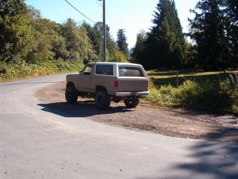 1985 Ford Bronco - The rear passenger side of a lifted, tan 1980s Ford Bronco is parked on a gravel shoulder next to a paved road. Tall green trees and a clea…