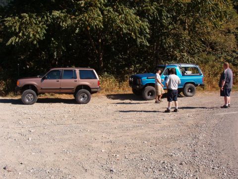 1985 Ford Bronco - Two lifted off-road vehicles, a brown SUV and a blue Bronco, are parked on a gravel road next to a wooded area. Three men stand near the bl…