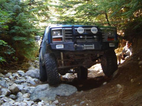 1985 Ford Bronco - A blue Ford Bronco is navigating a rocky, wooded trail. The front of the vehicle is visible, equipped with off-road lights and a winch.