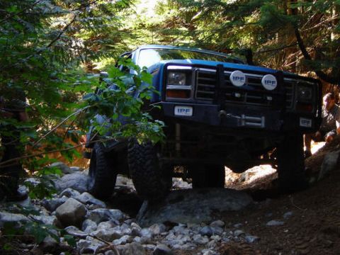 1985 Ford Bronco - A blue 1985 Ford Bronco is navigating a rocky, wooded trail. The front of the vehicle is visible, featuring IPF lights and a winch.