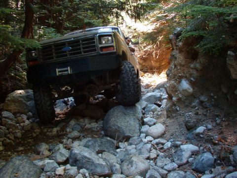 1985 Ford Bronco - A 1980s Ford Bronco navigates a rocky, wooded trail with large boulders and dense foliage. The front of the off-road vehicle, with its cust…
