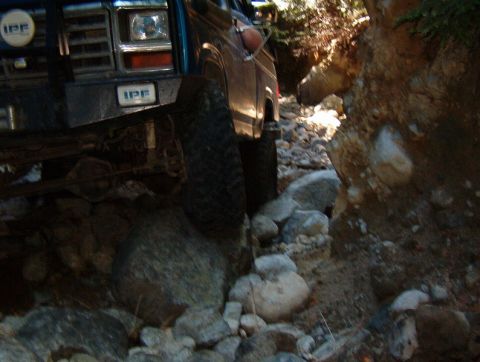 1985 Ford Bronco - Close-up, low-angle shot of the front driver's side of a dark blue SUV driving over a rocky, narrow trail. The vehicle's large tire is posi…