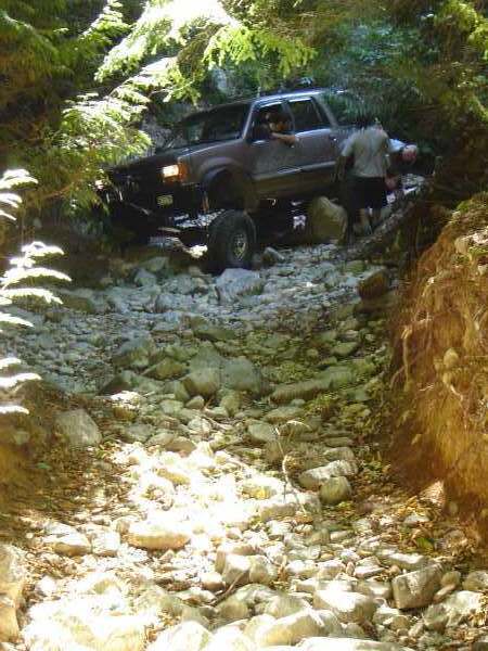 1985 Ford Bronco - A lifted, dark-colored pickup truck navigates a rocky, wooded trail. The driver is visible behind the wheel, and another person stands near…