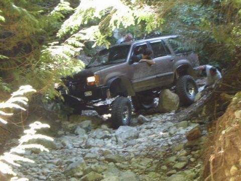 1985 Ford Bronco - A lifted, dark-colored SUV navigates a rocky, wooded trail with a person leaning out the driver's window. Another person is visible outside…