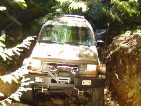 1985 Ford Bronco - A brown Ford Bronco is navigating a narrow, wooded trail with steep dirt banks on either side. The vehicle is equipped with a winch on the…