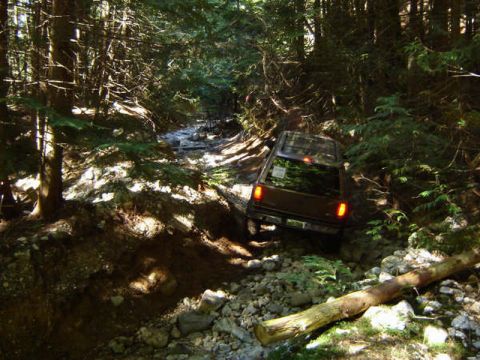 1985 Ford Bronco - A dark brown Ford Bronco is navigating a rocky, wooded trail, with its taillights illuminated. The vehicle is positioned at an angle, ascen…