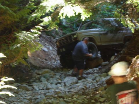 1985 Ford Bronco - A man is working on the tire of a light-colored Ford Bronco off-roading on a rocky, wooded trail.