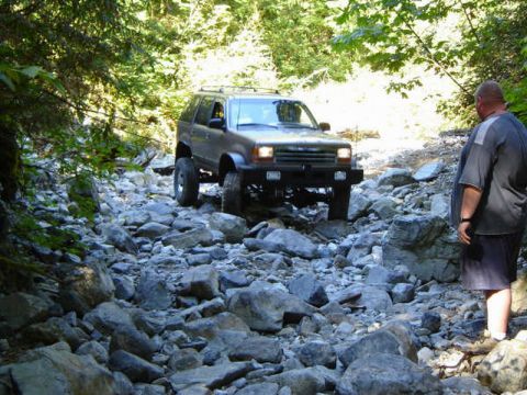 1985 Ford Bronco - A silver SUV navigates a rocky, wooded trail. A man stands to the right of the vehicle, observing the path.
