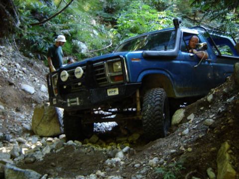 1985 Ford Bronco - A blue 1980s Ford Bronco is navigating a rocky, wooded trail with two people visible inside and one standing outside near the front.