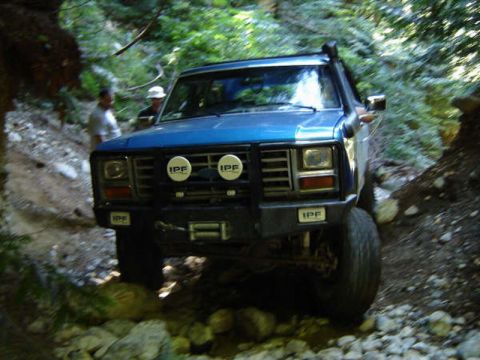 1985 Ford Bronco - A blue Ford Bronco is navigating a rocky, wooded trail. The front of the vehicle shows off-road lights and a snorkel.