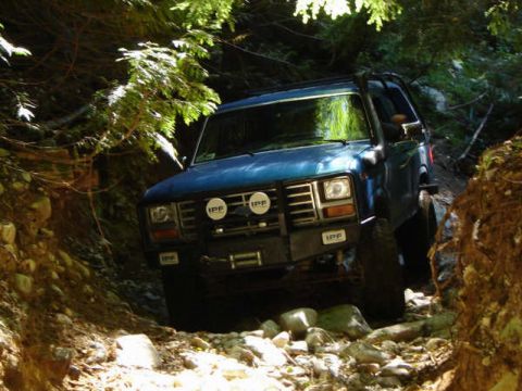 1985 Ford Bronco - A blue 1980s Ford Bronco navigates a rocky, wooded trail. The off-road vehicle features a winch and auxiliary lights on its front bumper.