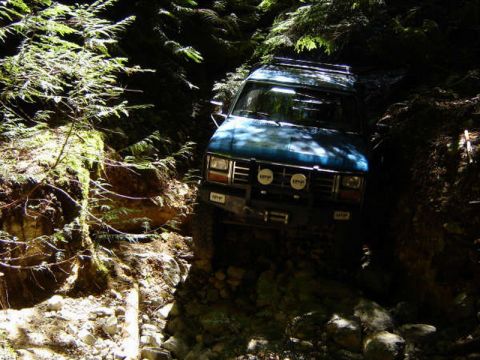 1985 Ford Bronco - A blue 1985 Ford Bronco is navigating a narrow, rocky trail surrounded by dense forest. The vehicle is equipped with auxiliary lights on th…