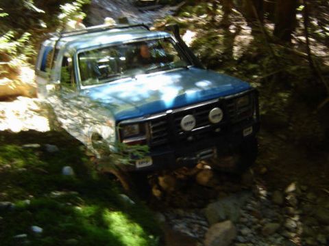 1985 Ford Bronco - A blue Ford Bronco is shown from a 3/4 front angle as it navigates a rocky, wooded trail. The vehicle has auxiliary lights mounted on the f…