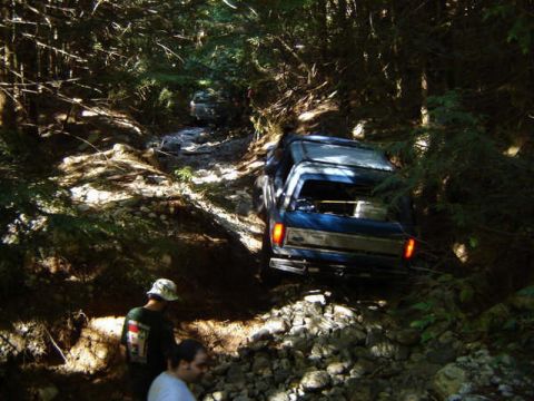 1985 Ford Bronco - A blue Ford pickup truck navigates a rocky, wooded trail. Two people are visible in the foreground, looking towards the truck.