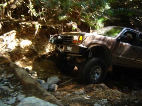 1985 Ford Bronco - A lifted, brown SUV is navigating a rocky, wooded trail with sunlight filtering through the trees. The vehicle's large tires and off-road m…