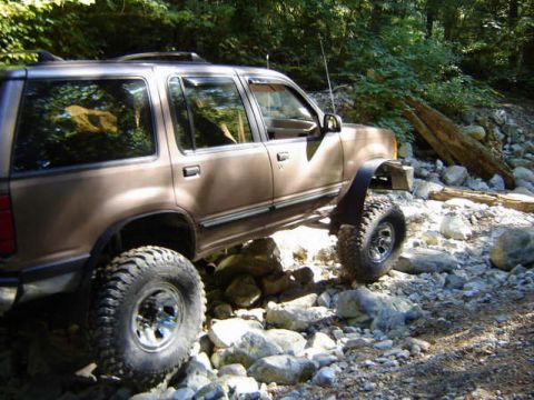 1985 Ford Bronco - A brown SUV is shown from the side, navigating a rocky, wooded terrain. The vehicle has large, off-road tires and appears to be lifted.