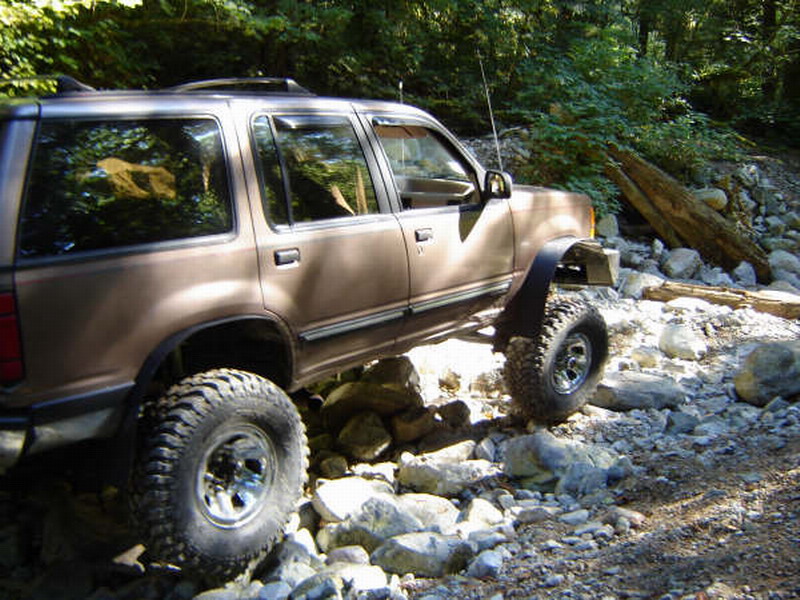 Brown SUV with large off-road tires on a rocky trail in a forest.