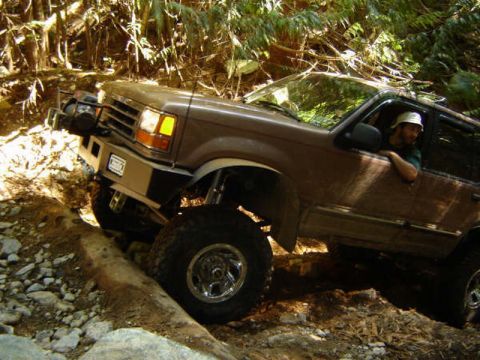 1985 Ford Bronco - A brown Ford Explorer is navigating a steep, rocky trail with a large log in the foreground. The driver, wearing a hat, is visible through…
