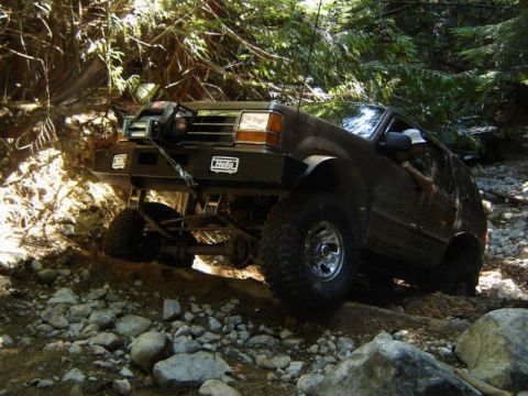 1985 Ford Bronco - A dark-colored, lifted SUV with large off-road tires navigates a rocky, wooded trail. The vehicle features a winch and aftermarket bumper w…