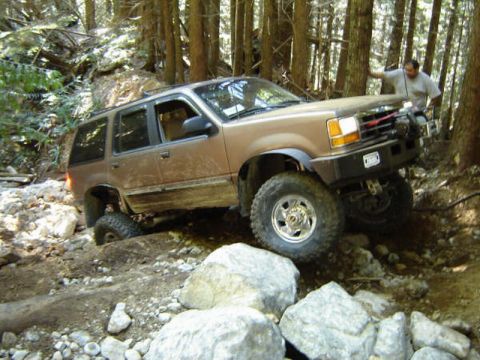 1985 Ford Bronco - A tan Ford Explorer is navigating a rocky, wooded trail with its front tire significantly elevated. A man stands near the front of the vehi…