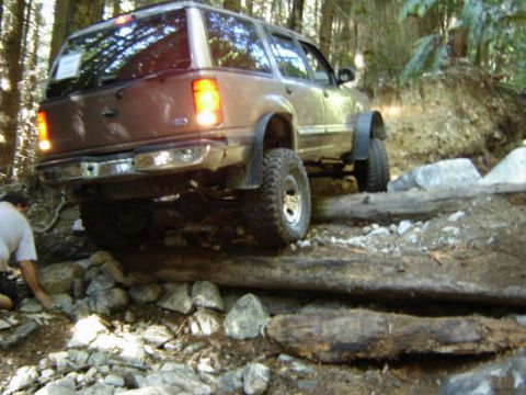 1985 Ford Bronco - A brown SUV is navigating a rough, wooded terrain, driving over large logs and rocks. The rear of the vehicle is visible, showing its taill…