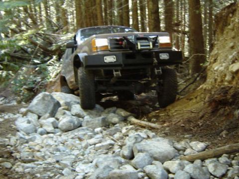 1985 Ford Bronco - A brown off-road vehicle navigates a rocky, wooded trail, with its front end prominently featured as it climbs over large stones. The vehic…