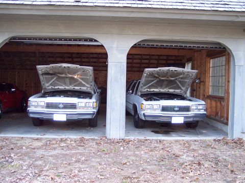 2001 Ford Crown Victoria - Two white Ford Crown Victorias are parked side-by-side in a garage with their hoods open, revealing their engines.
