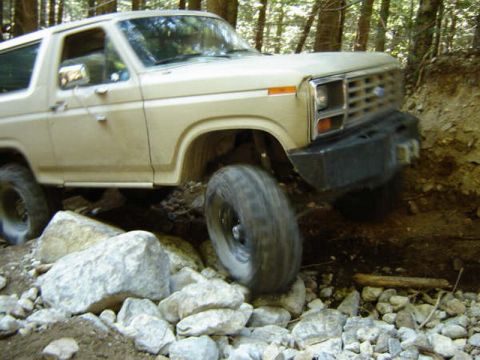 1985 Ford Bronco - A tan 1980s Ford Bronco is navigating a rocky, wooded trail, with its front tire on a large rock and its front bumper close to a dirt emban…